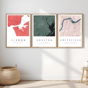 May include: Three framed city map prints. The maps are of Lisbon, Houston, and Louisville. The Lisbon map is red, the Houston map is green, and the Louisville map is pink. Each map has the city name and coordinates printed below.