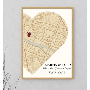 May include: A framed print featuring a heart-shaped map of a city, with a red heart-shaped marker. The print includes the names "MARTIN & LAURA" and the text "Where Our Journey Began" along with coordinates. The frame is a light brown color.