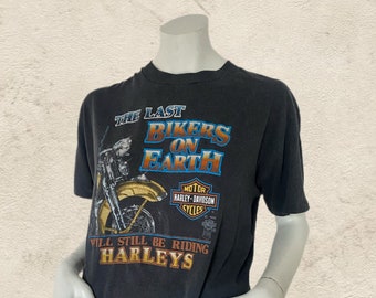 May include: A black Harley-Davidson t-shirt with the text "THE LAST BIKERS ON EARTH" and a motorcycle graphic. The shirt also features the Harley-Davidson Cycles logo. The shirt is displayed on a mannequin.