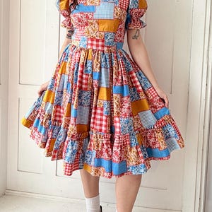 Könnte beinhalten: Ein farbenfrohes Patchwork-Kleid mit kurzen, gepufften Ärmeln und einem ausgestellten Rock. Das Kleid hat eine Mischung aus floralen, karierten und einfarbigen Stoffquadraten in Rot, Blau, Gelb und Weiß. Das Kleid ist knielang und hat ein figurbetontes Oberteil.