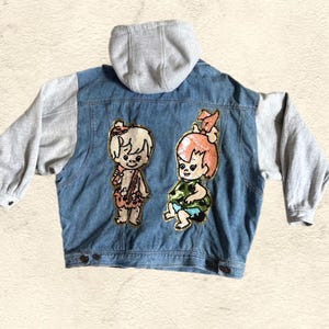 May include: A blue denim jacket with a gray hooded sweatshirt lining. The jacket has two embroidered patches of cartoon characters, a boy and a girl, on the back.