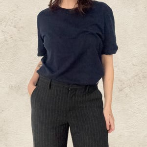 May include: A navy blue t-shirt and black pinstripe trousers. The t-shirt has short sleeves and a crew neck. The trousers are high-waisted with vertical stripes. The outfit is simple and classic.