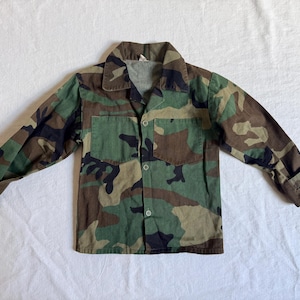 May include: A camouflage-patterned button-up shirt with long sleeves. The shirt features a classic collar, two chest pockets, and buttoned cuffs. The fabric displays a mix of green, brown, and black hues.