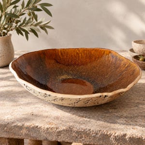 May include: A shallow, wide bowl with an irregular, organic rim. The interior features a glossy, deep amber and brown glaze with a speckled finish, while the exterior has a textured, matte cream-colored surface with dark, pitted detailing.