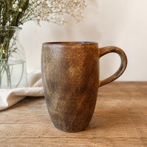May include: A tall, cylindrical ceramic mug with a textured, mottled brown and bronze finish. The mug features a curved handle on the right side and a smooth, slightly tapered base. The surface has a weathered, earthy appearance with dark brown tones.