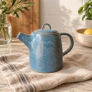 May include: A cylindrical ceramic teapot in a speckled blue glaze with a matching lid featuring a small arched handle. The teapot has a short, angled spout and a curved side handle, showcasing a textured finish with dark brown speckles.