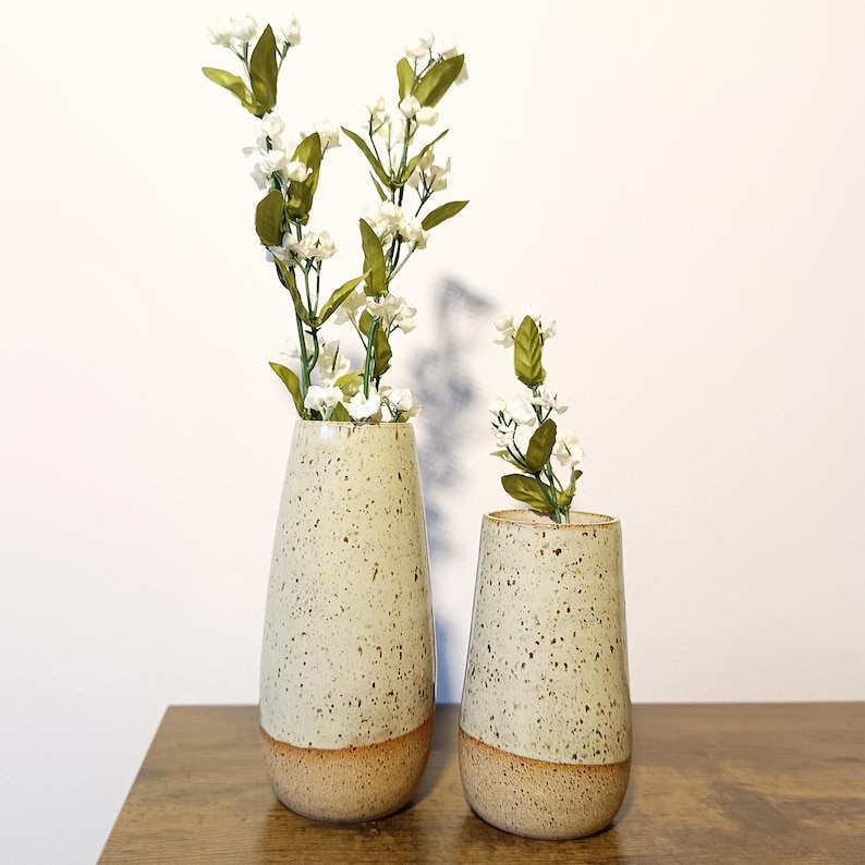 K&ouml;nnte beinhalten: Zwei gesprenkelte Keramikvasen, eine hohe und eine niedrige, mit beigefarbenem Sockel. Beide Vasen sind mit wei&szlig;en Blumen und gr&uuml;nen Bl&auml;ttern gef&uuml;llt. Die neutralen Farben passen in jedes Zuhause.