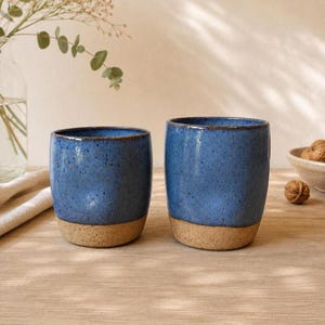 May include: Two cylindrical ceramic cups with a speckled blue glaze on the upper portion and a natural, unglazed tan base. The cups have a smooth, rounded rim and a slightly tapered shape, showcasing a contrast between the textured clay and the finish.