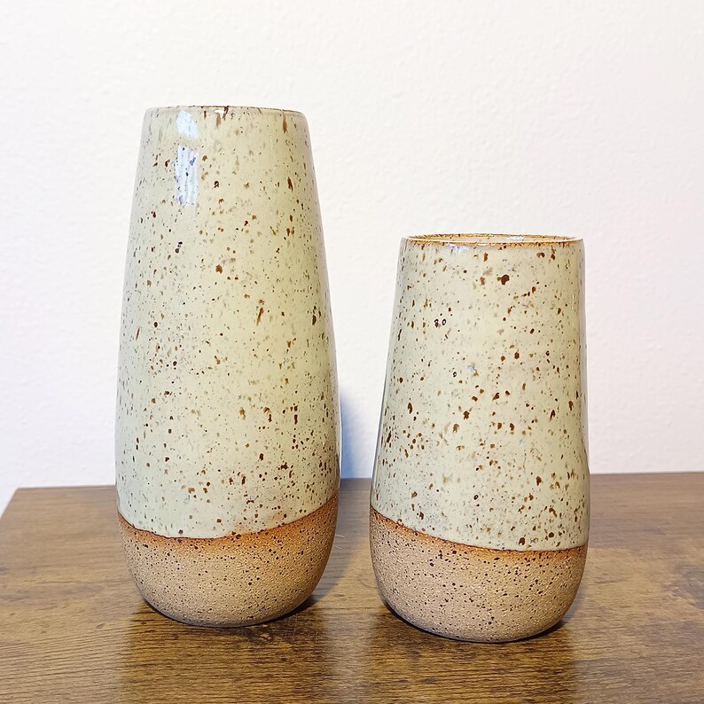 K&ouml;nnte beinhalten: Zwei gesprenkelte Keramikvasen. Die gr&ouml;&szlig;ere Vase ist etwa 25 cm hoch, und die kleinere Vase ist etwa 18 cm hoch. Beide haben eine hellgr&uuml;ne, gesprenkelte Glasur &uuml;ber einem beigen Sockel. Die Vasen haben ein modernes, minimalistisches Design.