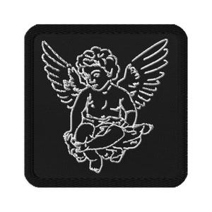 May include: Embroidered patch featuring a white cherub design on a black square background. The cherub is depicted with wings and is seated. The patch has a stitched border.