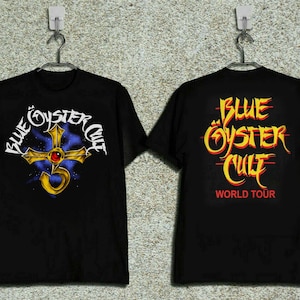 May include: Black t-shirts featuring the band Blue Öyster Cult. The front design includes the band's name and a gold cross with a red gem. The back of the shirt displays the band's name and "World Tour" in red.