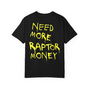 May include: Black t-shirt featuring the yellow text "NEED MORE RAPTOR MONEY". The text is in a dripping style. The shirt has a crew neck and short sleeves.