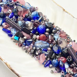Può includere: Una collezione di perline assortite nelle tonalità del blu, viola e rosa, con varie forme tra cui pezzi rotondi, sfaccettati, cilindrici e a forma di farfalla, disposti in un denso gruppo su una superficie bianca con un bordo dorato.