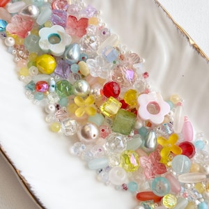 Può includere: Una collezione di perline e abbellimenti colorati di varie forme e dimensioni, tra cui fiori, farfalle e perle. Le perline sono disposte su un piatto bianco con bordo dorato, mostrando una varietà di colori e texture, perfette per progetti creativi.