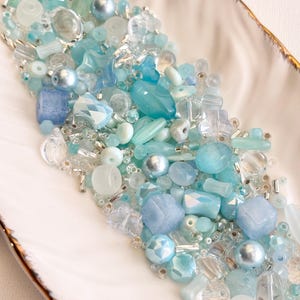 May include: A collection of various blue and clear beads in a white, leaf-shaped dish with a gold rim. The beads include round, faceted, and irregular shapes in shades of light blue, turquoise, and clear, with some silver accents.