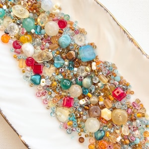 Può includere: Una collezione di perline colorate di varie forme e dimensioni, tra cui forme rotonde, quadrate e irregolari. Le perline sono in tonalità di blu, verde, rosso, giallo e marrone. Sono disposte su un piatto bianco, smerlato con un bordo dorato.