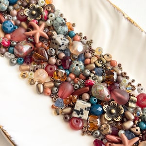 May include: A selection of colourful beads, including various shapes and sizes, such as round, teardrop, and cube, are arranged on a white, leaf-shaped dish with a gold rim. The beads are in shades of blue, red, brown, and gold, with some featuring floral and starfish accents.