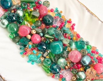 May include: A collection of loose craft beads in various shades of green, teal, and pink. The assortment includes faceted, round, and butterfly-shaped beads in glass, plastic, and pearl-like finishes, displayed in a long, white, curved dish.