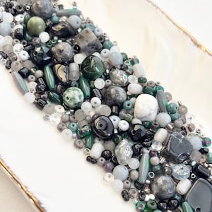 May include: A collection of beads in various shapes and sizes, including round, oval, and faceted, in shades of green, gray, black, and white. The beads are arranged on a white, oblong dish with a gold rim. The beads are made of different materials.