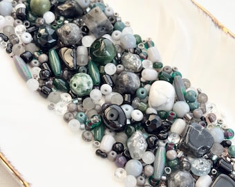 May include: A collection of beads in various shapes and sizes, including round, oval, and faceted, in shades of green, gray, black, and white. The beads are arranged on a white, oblong dish with a gold rim. The beads are made of different materials.