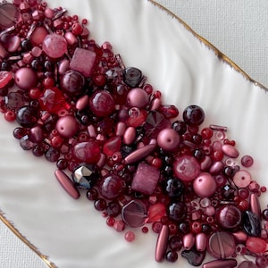 May include: A collection of assorted beads in various shades of red, burgundy, and maroon. The mix includes round, faceted, cube, and cylindrical shapes with matte, glossy, and translucent finishes, all arranged in a cluster on a white surface.