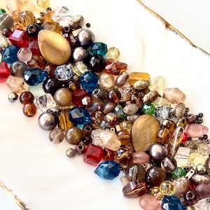 May include: A collection of assorted loose beads in various shapes, sizes, and colors including deep blue, amber, red, clear, and metallic grey. The beads feature faceted, smooth, and oval textures, with two prominent smooth tan oval stones.