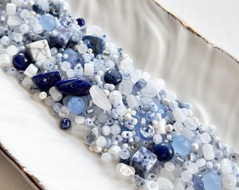 May include: A selection of blue and white beads in a variety of shapes and sizes, including round, square, and irregular, are displayed in a white, leaf-shaped dish with a gold rim. The beads vary in shades from dark navy to light sky blue.