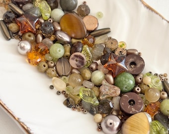 May include: A collection of assorted beads in various shapes, sizes, and colors, including brown, green, and clear. The beads are arranged on a white rectangular dish with a gold rim. The beads include round, oval, and star-shaped elements.