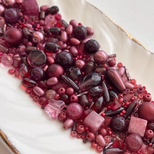 May include: A collection of various red and pink beads in different shapes and sizes, including round, oval, and faceted, are arranged on a white, scalloped dish with a gold rim. The beads range in color from light pink to deep burgundy.