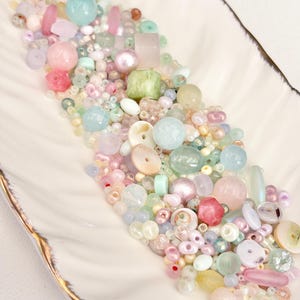 May include: A collection of colorful beads in various shapes and sizes, including round, cube, and irregular forms. The beads are in shades of pink, blue, green, and yellow, with some iridescent and translucent elements. The beads are displayed in a white, feather-shaped dish.