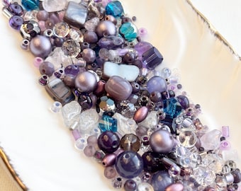 May include: An assortment of purple, lavender, and clear beads in various shapes and sizes. The beads are displayed in a white, feather-shaped dish with a gold rim. The beads include round, faceted, and irregular shapes.