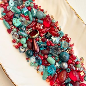 Puede incluir: Una colección de cuentas coloridas de varias formas y tamaños, incluyendo redondas, facetadas y en forma de corazón, en tonos rojos, turquesas y dorados. Las cuentas están dispuestas en una fuente blanca oblonga con borde dorado, para bisutería.