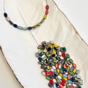 May include: A gold-coloured necklace featuring a rainbow of oval beads. A collection of similar beads in various colours rests on a white, feather-shaped dish with gold trim. The beads have gold-coloured edges.