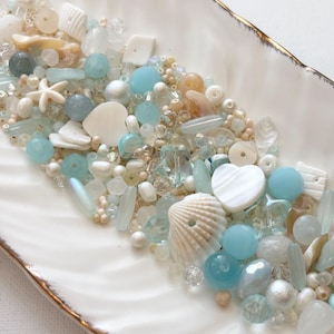 May include: A collection of assorted beads in shades of light blue, white, and cream. The mix includes round, faceted, and heart-shaped beads, along with small white starfish and seashell-shaped charms, all presented in a white dish.
