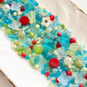 May include: A vibrant assortment of beads in shades of blue, green, red, and white, displayed on a white dish with a gold rim. The beads vary in shape, including round, flower, and faceted designs. These beads are likely intended for jewellery making or crafting projects.