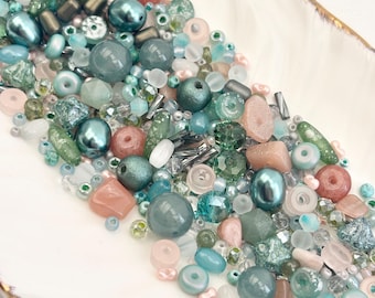 May include: A collection of beads in various shapes and sizes, including round, oval, and irregular, in shades of teal, pink, and clear. The beads are arranged in a white, leaf-shaped dish with a gold rim. The beads are likely for jewelry making.