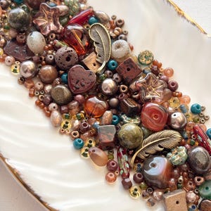 May include: A collection of assorted craft beads in various shapes, sizes, and textures, including metallic feather charms, heart-shaped beads, star-shaped glass beads, and round stone-like beads in shades of brown, bronze, copper, and teal.