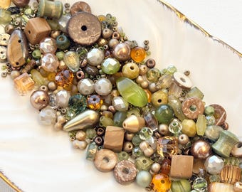 May include: A collection of assorted beads in shades of olive green, bronze, brown, and gold. The mix includes various shapes such as spheres, cubes, cylinders, and faceted crystals, along with flat circular discs and teardrop-shaped pieces.