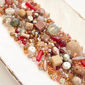 May include: A collection of assorted beads in various shapes, sizes, and colors, including red, brown, gold, and pearl. The beads are arranged in a white, rectangular dish with a gold rim. The beads are a mix of glass, stone, and other materials.
