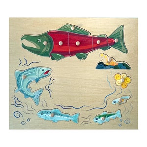 May include: Wooden puzzle depicting a salmon and its life cycle, with removable pieces. The salmon is red and green, and other fish are also shown. The puzzle is set against a light wood grain background, suitable for educational play.
