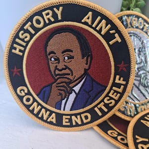 May include: Embroidered patch with the text "HISTORY AIN'T GONNA END ITSELF" encircling a portrait of a man in a suit. The patch has a black border with gold stitching and a red background.