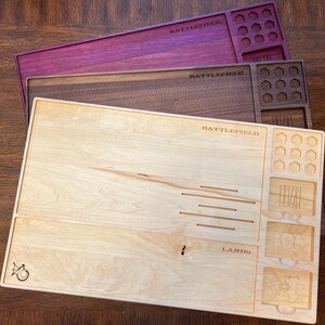 May include: Three wooden game trays in shades of purple, brown, and light wood. Each tray is rectangular with carved compartments and the word "BATTLEFIELD" etched across the top. The light wood tray has the word "LANDS" etched on it.