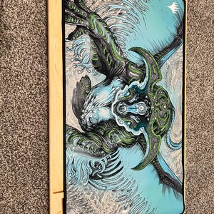 May include: A rectangular gaming mat featuring a detailed illustration of a creature with green and blue accents. The artwork is set against a light blue and gray background. The mat is bordered by a black trim and rests on a wooden frame.