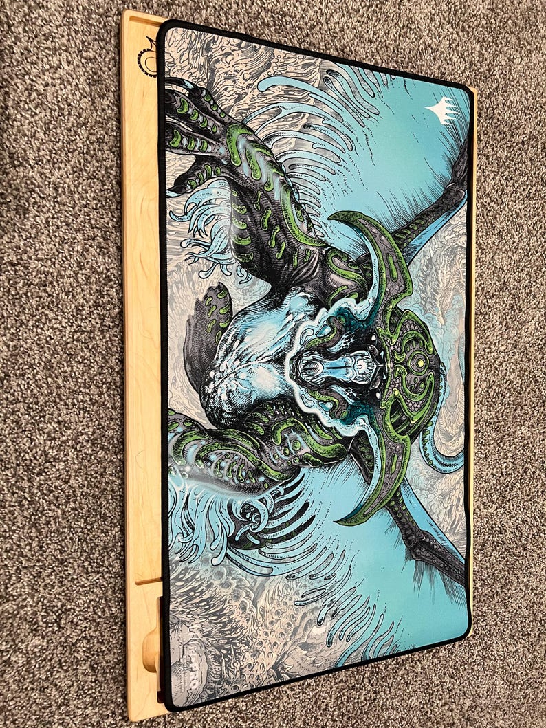 May include: A rectangular gaming mat featuring a detailed illustration of a creature with green and blue accents. The artwork is set against a light blue and gray background. The mat is bordered by a black trim and rests on a wooden frame.