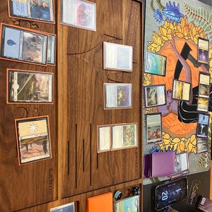 May include: A wooden game board with card slots and a play mat. Various Magic: The Gathering cards are arranged on the board. An orange card holder and dice are also visible. The word "LANDS" is printed on the board.