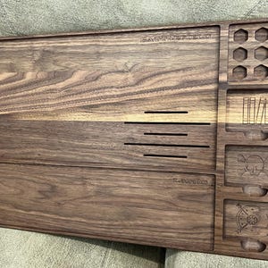 May include: A wooden tabletop organizer with multiple compartments and carved details. The organizer features the word "BATTLEFIELD" and "LAIR" carved into the wood. It includes various shaped storage areas, including hexagonal and rectangular slots, and a skull and crossbones design.