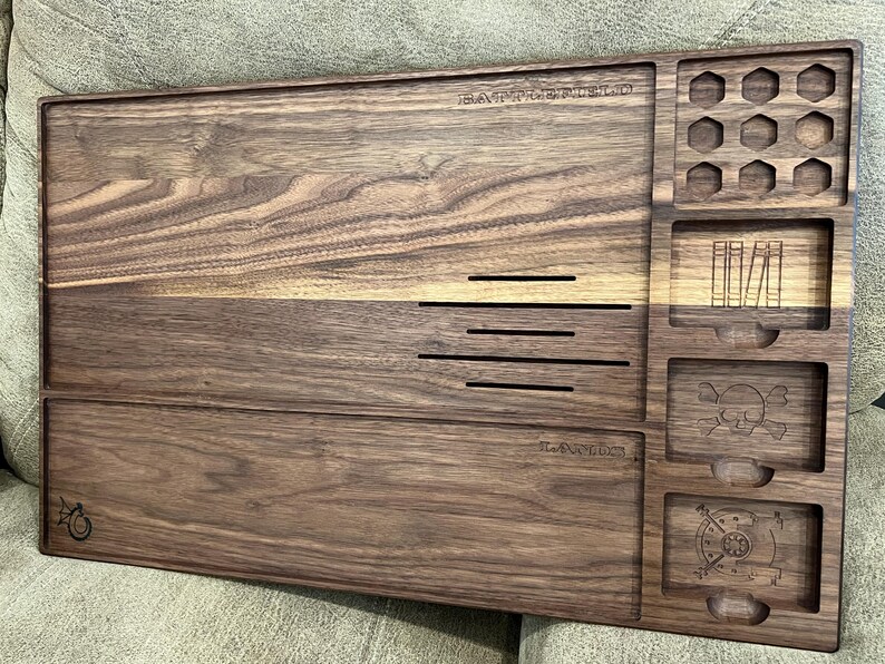 May include: A wooden tabletop organizer with multiple compartments and carved details. The organizer features the word "BATTLEFIELD" and "LAIR" carved into the wood. It includes various shaped storage areas, including hexagonal and rectangular slots, and a skull and crossbones design.