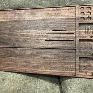 May include: A dark brown wooden tray with multiple compartments and etched designs. The tray features slots, a skull and crossbones, and the text "BATTLESTATION.ID" and "The Awesome's". Ideal for organizing small items.