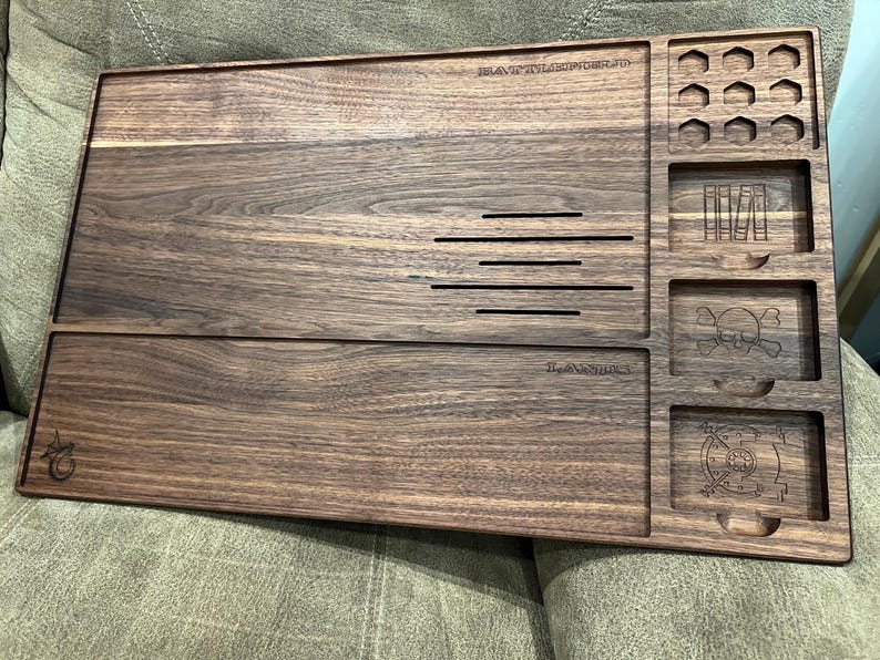 May include: A dark brown wooden tray with multiple compartments and etched designs. The tray features slots, a skull and crossbones, and the text "BATTLESTATION.ID" and "The Awesome's". Ideal for organizing small items.