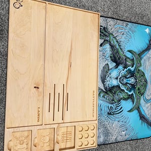 May include: A light-colored wooden game board with compartments and etched designs. The board has slots for cards, dice, and tokens, with the word "LANDS" and "BATTLEFIELD" etched on it. A blue and green graphic mat is next to the board.
